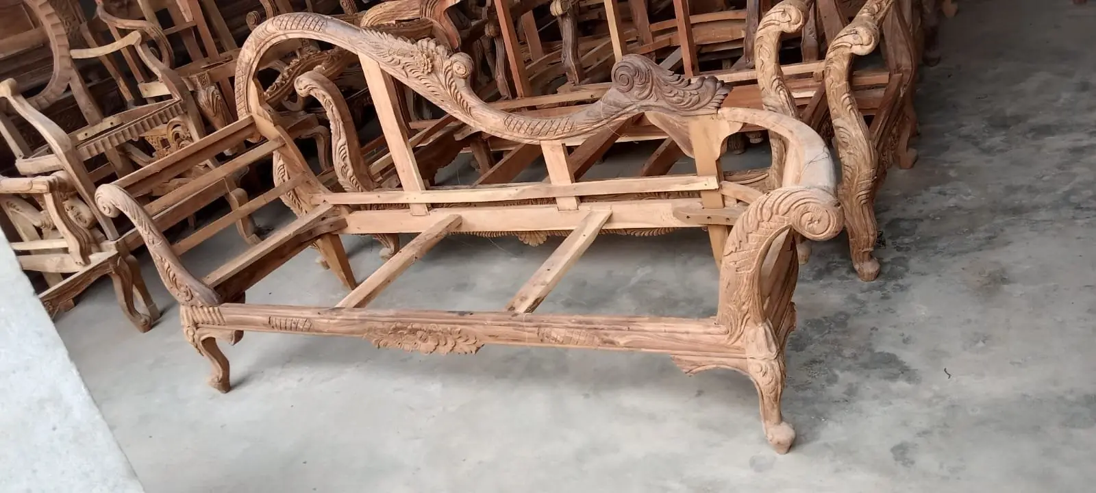 An unfinished Floral Hand Carved Sheesham Wood Dewan frame sits in a workshop, showcasing intricate woodwork and elegant curved armrests.