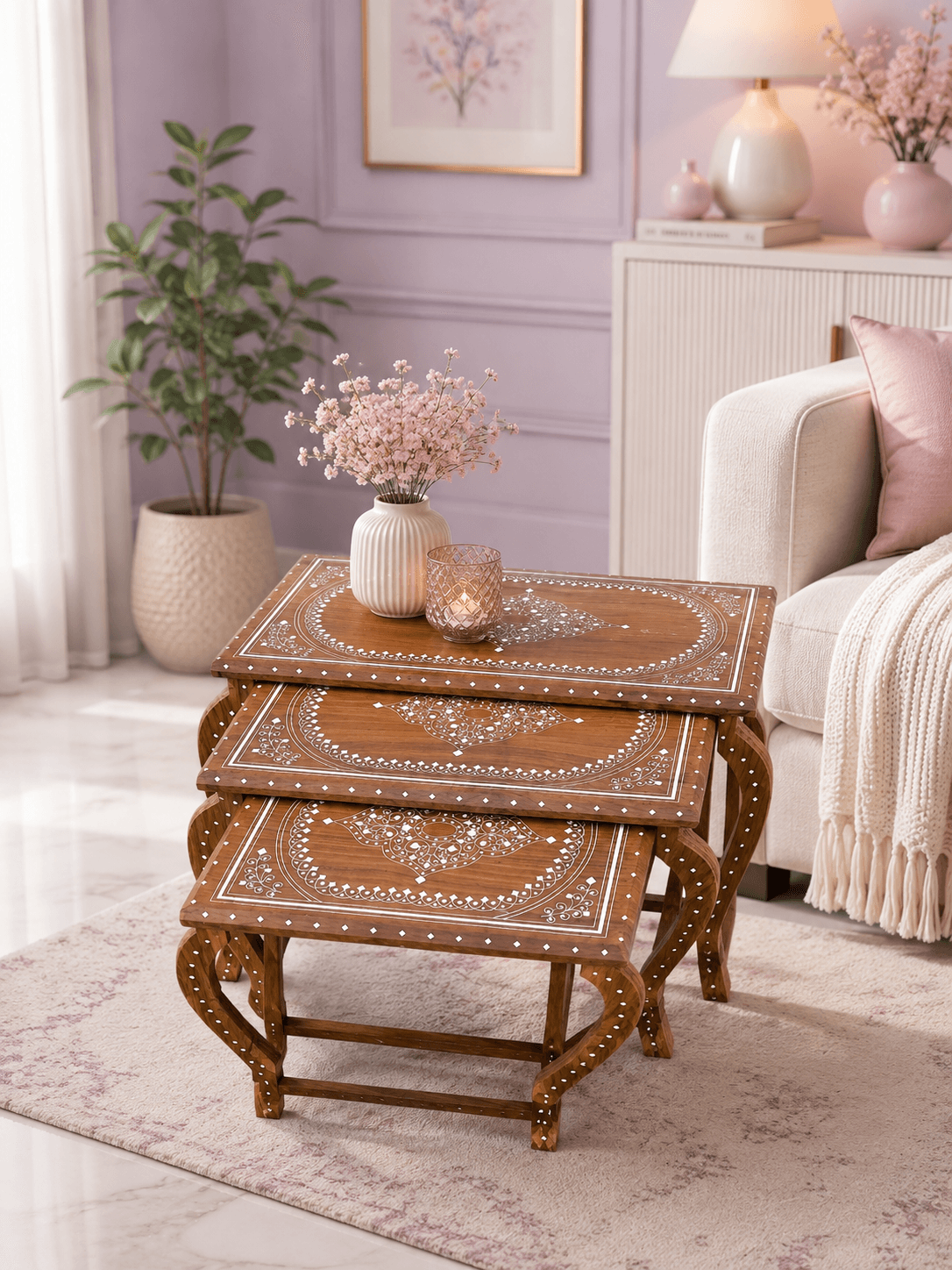 A set of three nested wooden tables with intricate white floral patterns, placed on a rug in a cozy living room with soft pink wall decor.