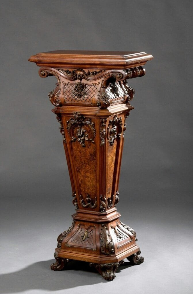 An ornate antique wooden pedestal with intricate carvings and burl wood panels, standing against a plain gray background for our About Us page.