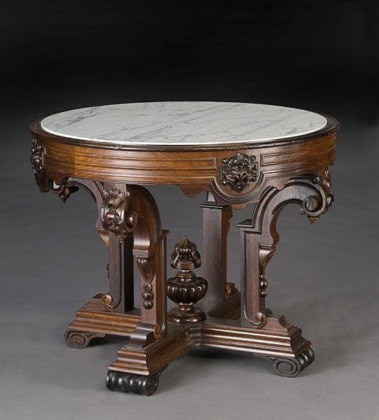 An antique round wooden table with a white marble top, featuring ornate carved legs and a central finial, set against a dark gray background.