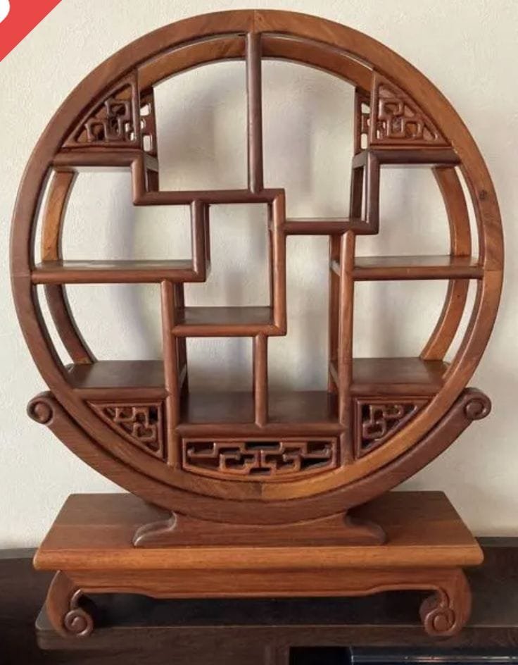 A circular wooden display shelf with intricate, staggered compartments, resting on a rectangular base against a plain white wall in an About Us view.