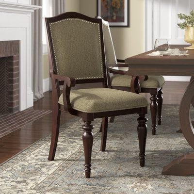 A traditional dining chair with armrest featuring dark wood legs and beige fabric upholstery, placed in a cozy dining room with a fireplace.