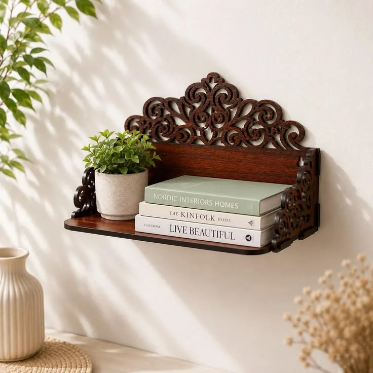 A dark wooden wall shelf with ornate scrollwork holds a small potted green plant and a stack of three books against a plain white wall.