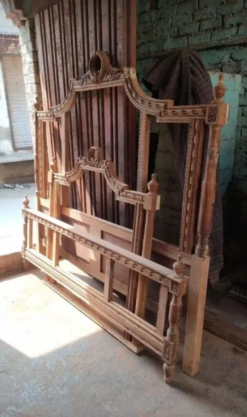 An unfinished wooden bed frame with intricate carved details leaning against a textured wall in a workshop, reflecting Cladsi craftsmanship.