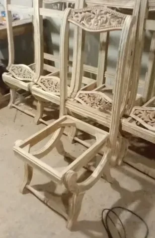 Several unfinished wooden chairs with intricate carvings stand in a workshop, showcasing the craftsmanship typical of Cladsi furniture production.