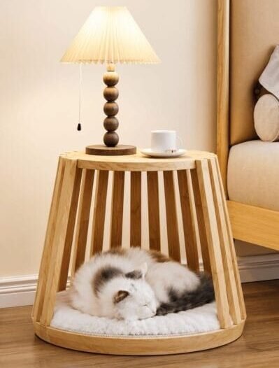 A fluffy cat sleeps inside a wooden slatted Cladsi side table, which is topped with a warm lamp and a coffee mug next to a bed in a cozy bedroom.